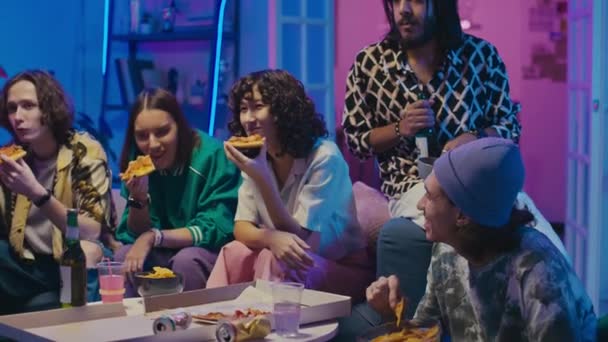 六个快乐的年轻人坐在电视机前的沙发上 在家里用蓝色 粉色霓虹灯照明 喝啤酒 吃外卖披萨 看电影或系列片 拍了一张中等比例的平底照 — 图库视频影像