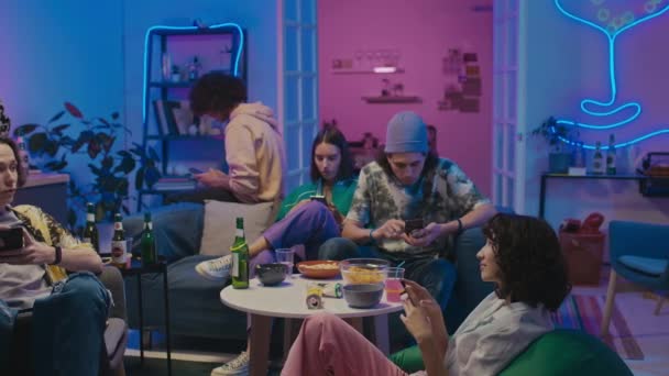 满满一盘疲惫的年轻男女学生坐在房间里 家庭聚会后都点着蓝色和粉色的霓虹灯 桌上放着空啤酒瓶 他们都在忙着用智能手机 — 图库视频影像