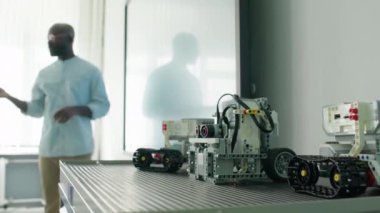Plastik yapı tuğlalarından yapılmış küçük oyuncak robotların yakın çekim odak çekimi ve beyaz tahtada duran siyah erkek öğretmen ve bulanık arka planda teoriyi açıklayan