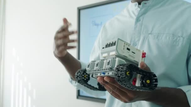 メガネで若い黒人男性教師の手と顔のミディアムクローズアップタイルショット 小さなトラクターロボットモデルを保持し ロボットレッスン中にクラスに話す — ストック動画