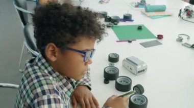 Orta açılı, kıvırcık saçlı, gözlüklü, robotik derste masasında oturan, kağıda tasarım çizen ve arka planda robot modeller yapan Afro-Amerikan bir çocuğun orta açı çekimi.