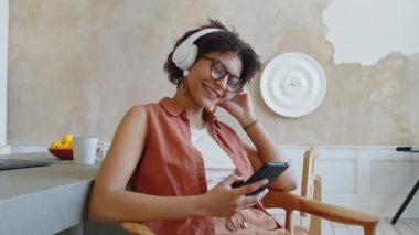 Çok ırklı, koyu tenli, kablosuz kulaklıklı bir kadının son moda minimalist tasarımıyla evde tek başına oturup müzik dinlerken, gülümserken ve akıllı telefondan selfie çekerken orta boy bir fotoğrafı.