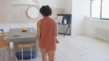 Orta boy siyah bir kadın dizüstü bilgisayarla şık, minimalist çağdaş bir dairede yürüyor, masada oturuyor, not defteri alıyor ve klavyede daktilo kullanıyor.