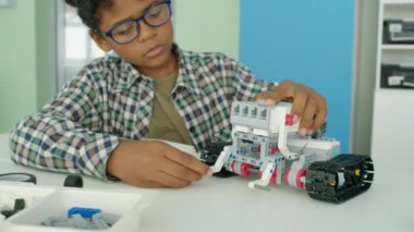 9 yaşındaki inek, gözlüklü ve ekose gömlekli Afro-Amerikan bir çocuğun okul sonrası hobi kulübünde otururken ve plastik bloklardan oyuncak robot yaparken orta ölçekli yakın çekimi.