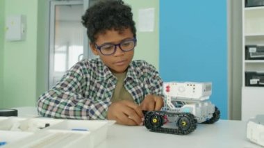 Orta boy, gözlüklü, kıvırcık saçlı, plastik bloklardan oyuncak robot traktör yapan ve masaya iten Afrikalı Amerikalı bir çocuğun orta boy fotoğrafı.