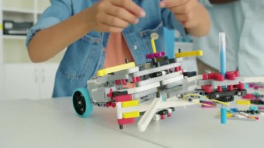 Robotik kulübündeki karmaşık boyutsal yapının tepesine parlak plastik oyuncaklar koyan tanınmayan genç bir çocuğun yakın plan fotoğrafları ve arka planda koyu tenli bir öğretmen.