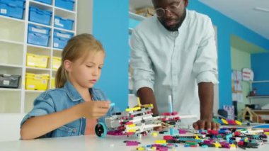 Okul sonrası robotik kulübünde oturan ve plastik bloklardan oyuncak modeller yapan orta boy sarışın beyaz bir kız ve siyah erkek öğretmen izlenip önerilerde bulunuyor.