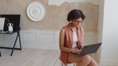 Orta halli, gözlüklü, şık minimalist oturma odasında taburede oturan, alçı heykeltraş sanatı ve bej duvarlı, dizüstü bilgisayarda yazan ve pencereye bakan çok kültürlü genç bir kadının fotoğrafı.