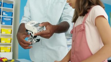Siyahi erkek öğretmenin robot modelin bir parçasını elektrikli motorla tutarken ve okul sonrası hobi kulübünde genç beyaz bir kız öğrenci için inşaatını gösterirken orta boy yakın çekim çekimi.