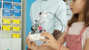Elektrikli robotun bir parçasını elinde tutarak onu izleyen ve montajı için tavsiye veren Afrikalı Amerikalı erkek öğretmenle konuşan inek beyaz kızın orta boy yakın çekimi.
