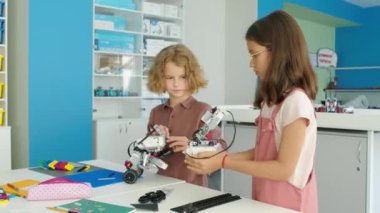 Orta boy beyaz bir erkek ve kız çocuğu okul sonrası bilim kulübünde oturmuş plastik oyuncak inşaat setlerinden elektrikli robot modeller yapıyor ve konuşuyorlar.
