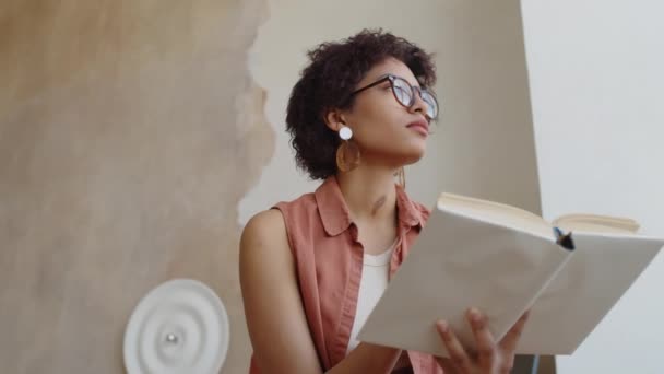自宅でトレンディなベージュウォールで座って 読書本で夢のような若い黒人女性のミディアムローアングルショット — ストック動画