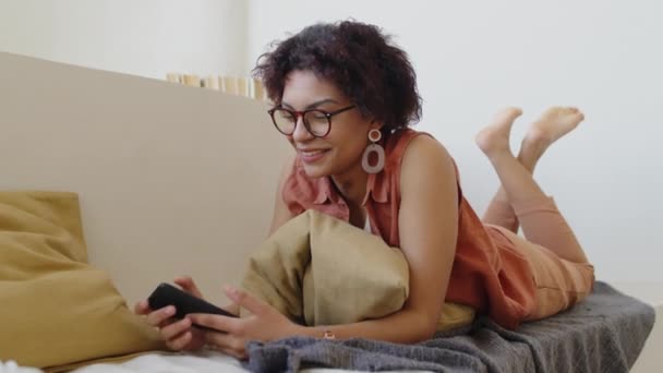 メガネの若い多民族女性のフルショット 短い波状の髪 ソファに胃の上に横たわる袖なしのシャツ スマートフォンを保持 ソーシャルネットワーク上のフィードをスクロールし — ストック動画
