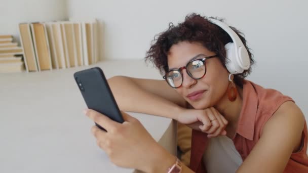 若い女性のクローズアップショット カーリーヘア メガネ 大きな白いワイヤレスヘッドフォン 顎の下に腕で座って スマートフォンや笑顔でメッセージをスクロール — ストック動画