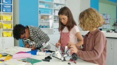Orta boy beyaz ve Afrikalı Amerikalı çocukların masada durup, okul sonrası kulüplerinde plastik inşaat setlerinden robot modeller yapıp sohbet etmelerinin fotoğrafı.
