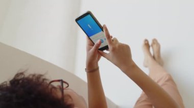 Beyaz duvara yaslanmış, ayakları üzerinde uzanmış, akıllı telefon tutan ve ses kitabı mobil uygulamasındaki oynatma tuşuna basan kadınların yakın çekim görüntüleri.