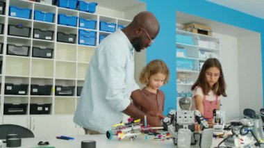 Açık plastik yapı taşları ve elektrikli robot modelleriyle çocuk hobisi kulübünde masa başında duran beyaz erkek ve kız çocuklarıyla dolu. Ve siyah erkek öğretmen onlara ders verme görevini anlatıyor.