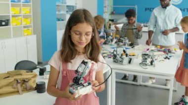 Okul sonrası hobi kulübünde elinde elektrikli robot modeliyle poz verip gülümseyen 9 yaşındaki beyaz bir kızın orta boy portresi. Duvardaki metin Kiril Robot Kuantum ile