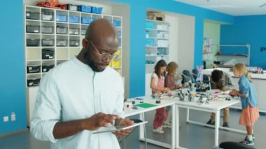 Orta boy Afrikalı Amerikalı bir adamın okuldan sonra robotlar kulübünde durup tablet bilgisayar kullanırken ve arka planda renkli plastik yapı taşlarından modeller yaparken görüntüsü.