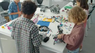 Okul sonrası masada elektrikli robot modeller yapan genç erkeklerin orta açı çekimi ve parlak plastik bloklardan yapılar inşa eden kızlar.