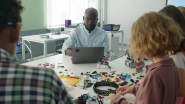 Okuldan sonraki fen dersinde, çeşitli çocuklar robot modeller yaparken, Afrikalı Amerikalı erkek öğretmenin dizüstü bilgisayarda çalışırken orta omuz üstü çekimleri.