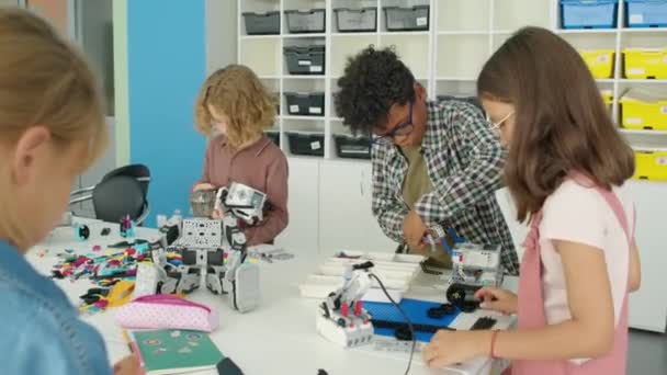 从放学后科学俱乐部明亮的塑料建筑套件上拍下的不同年龄的男孩和女孩在教室里围坐在桌旁制作电动机器人模型的照片 — 图库视频影像