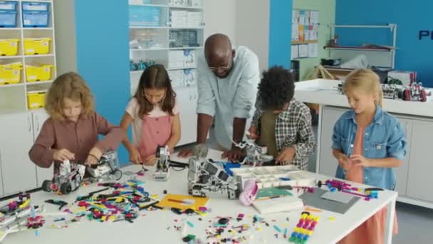 教室のテーブルの周りに立って プラスチック製の建設キットからロボットモデルや構造を構築し 黒人男性教師がアドバイス — ストック動画