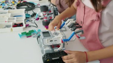 Genç bir kızın, gençlik robotik kulübünde masada dikilip, elektrikli robot modelinin çalışma tuşlarına basarken ve arka planda plastik inşaat bloklarıyla oynayan bir çocuğun yakın çekim görüntüleri.