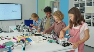 Hobi kulübünde masada yan yana duran, plastik inşaat setlerinden oyuncak robot modeller yapan ve sohbet eden dört mutlu çokkültürlü çocuğun orta boy fotoğrafı.