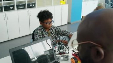 Okul sonrası kulüpte oyuncak robotlar için bilgisayar yazılımı yazan siyah erkek bilgisayar öğretmeninin orta omuzlu fotoğrafı ve arka planda dizüstü bilgisayarlar üzerinde çalışan genç çocuklar.