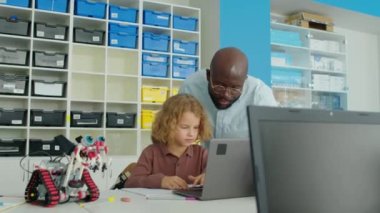 Kafkasyalı genç bir çocuğun robotlar kulübünde dizüstü bilgisayarla teknoloji projesi üzerinde çalışırken çekilmiş orta boy fotoğrafı. Afro-Amerikan öğretmen izliyor, açıklıyor ve hatayı düzeltiyor.