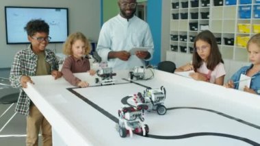 Bir grup genç, etnik çeşitliliğe sahip erkek ve kız ve zenci erkek öğretmen çocuk hobisi kulübünün etrafında toplanmış, elektrikli robotların hareketlerini izliyor, neşeyle gülüyor ve zıplıyorlar.