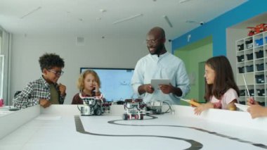 Bir grup ergen erkek ve kız çocuğu ve Afrika kökenli Amerikalı öğretmen çocuk teknoloji kulübünde masanın etrafında durup elektrikli robotların hareketlerini, dövüşlerini, gülüşmelerini, neşeyle zıplamalarını izliyorlar.