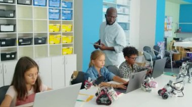 Orta boy Afrika kökenli Amerikalı erkek bilgisayar öğretmeninin sınıfta dolaşırken ve çeşitli genç öğrencilerin robotlar için dizüstü bilgisayarlara yazdıkları programları incelerken tam bir görüntüsü. Kiril RoboKuantum metni