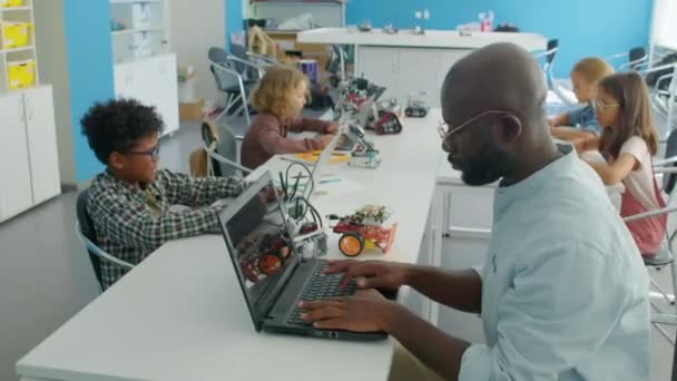 身着眼镜的非洲裔美国男性机器人老师和带着手提电脑坐在桌旁 编写电脑程式来操作电动机器人模型的中弧镜头 — 图库视频影像