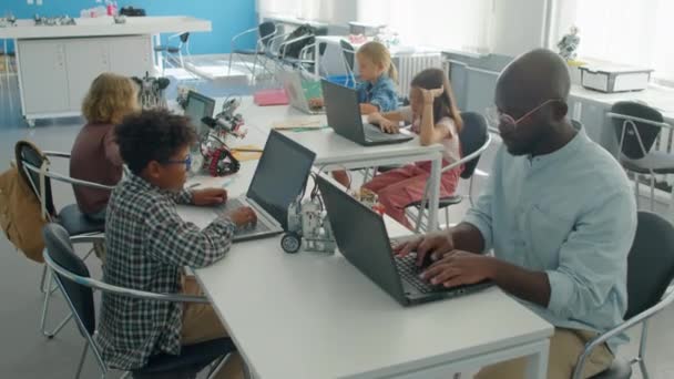 中等比例的黑人男教师和各种各样的男孩和女孩坐在信息技术业余爱好俱乐部的桌旁 编写计算机程序来操作和控制电动机器人模型 — 图库视频影像