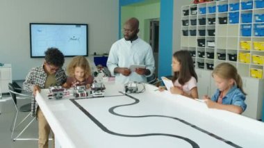 Bir grup genç ve Afrikalı Amerikalı öğretmen okul sonrası teknoloji kulübünde masanın etrafında dikilip elektro robot modelleri izleyip test ediyorlar.