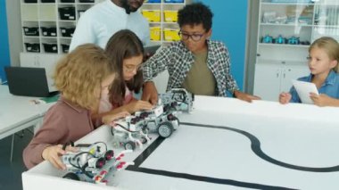 Heyecanlı dokuz yaşındaki çeşitli çocukların elektrikli robot modellerini çocuk hobileri kulübünde test ederken, kontrol panellerindeki düğmelere basarken ve siyahi erkek öğretmen onları izlerken ve rehberlik ederken orta boy çekimleri.