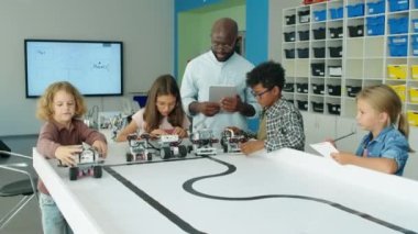 Heyecanlı çoklu etnik erkek ve kızların okuldan sonra robot modellerini fırlatıp neşeyle zıpladıkları ve gözlüklü siyahi erkek öğretmenlerin izleyip yardım ettiği bir sahne.