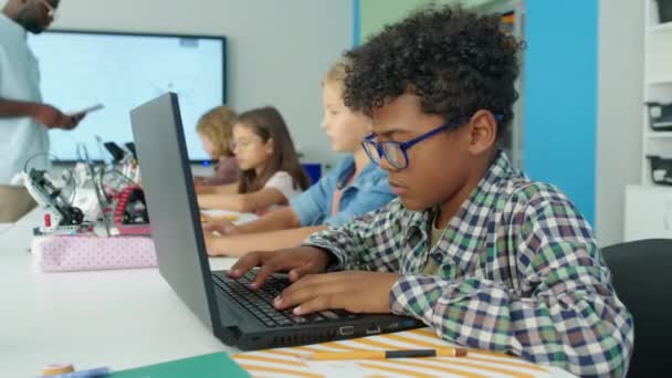 身着眼镜坐在机器人俱乐部桌旁的专注的非洲裔美国男孩的中侧镜头 以及以计算机为背景的电动机器人 同学和教师书写程序 — 图库视频影像
