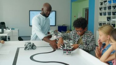 Gençlik robotik klübünde programlanabilir elektrikli robot modelleri izleyen bir grup orta sınıf öğrenci ve tablet bilgisayarlı siyah erkek bilişim öğretmeni.