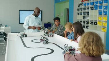 Okul sonrası teknoloji kulübünde dört farklı çocuğun elektrikli oyuncak robotlarla oynadığı orta boy fotoğraf ve tablet bilgisayarlı Afro-Amerikan erkek öğretmen.