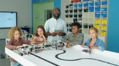 Afrika kökenli Amerikalı erkek teknoloji öğretmeninin orta boy portresi ve okuldan sonra robot modellerle poz veren ve gülümseyen dört farklı genç kız ve erkek çocuk.