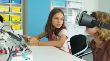Genç beyaz çocukların okul sonrası masada oturup konuştuğu tablet bilgisayarda kız sörfü yaptığı ve AR kaskı takıp sanal gerçeklik oyunu oynadığı orta ölçekli bir fotoğraf.