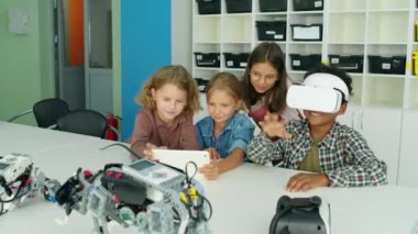 Modern çocuk yuvasında masada oturan, tablet bilgisayarda video ya da çizgi film izleyen, oynayan, VR kulaklık kullanan, gülümseyen ve gülen çocukların tam karesi.