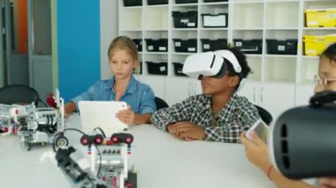 Gündüz kulübündeki masada oturmuş tablet bilgisayar kullanan orta boy çocuklar AR kulaklıklı Afro-Amerikan bir çocuk etrafa bakıyor ve izlenimlerini paylaşıyor.