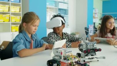 Kafkasyalı kızların tablet bilgisayarlı ve AR kulaklı Afro-Amerikan bir oğlanın gençlik robotları kulübünde masada oturup internette sanal gerçekliği keşfettiği orta boy bir fotoğraf.