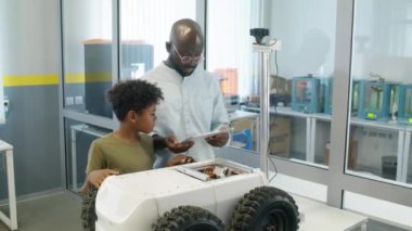 Afrika kökenli Amerikalı erkek mühendisin tablet bilgisayarla çektiği orta boy fotoğraf genç oğluna laboratuvarda dağıtım robotlarının kablolarını ve devrelerini gösteriyor.