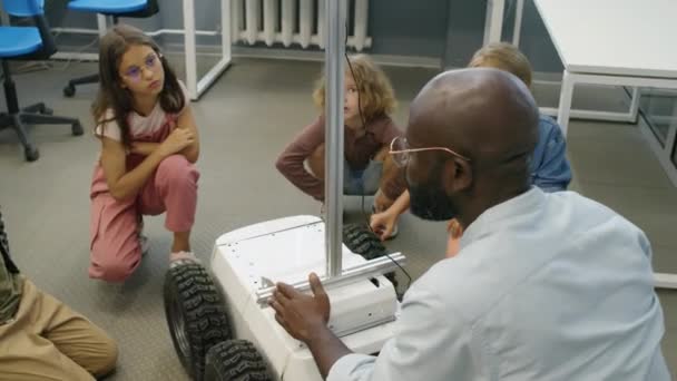教室里穿着休闲装的年轻多种族男孩和女孩围坐在送货机器人周围的中弧镜头 戴着眼镜的非洲裔美国男教师解释其设计和功能 — 图库视频影像
