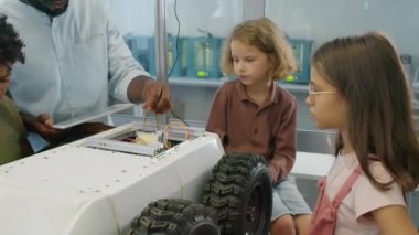 Afrika kökenli Amerikalı erkek teknoloji öğretmeninin, okul sonrası robotik klübünde ders sırasında çok ırklı genç erkek ve kız çocuklarına dağıtım robotlarının kablolarını ve devrelerini gösteren orta kavisli bir fotoğrafı.